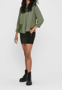 Vrouw in een olijfgroene losse blouse, zwarte suede minirok en zwarte veterlaarsjes, staand tegen een effen achtergrond.