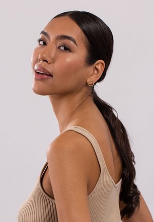 Model trägt ein beiges, geripptes Tanktop mit dünnen Trägern, das eine strukturierte Oberfläche aufweist. Kleine goldene Kreol Ohrringe betonen den Look.