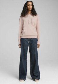 Sudadera rosa claro con bolsillo frontal, combinada con jeans de pierna ancha en denim oscuro y zapatillas ligeras. La tela parece suave y casual.