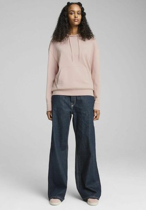 Lyserød hættetrøje med frontlomme, parret med meget brede mørke denim jeans og lyse sneakers. Stoffet ser blødt og afslappet ud.