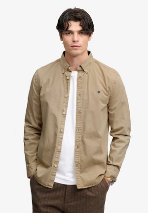 Ung mand med sort hår iført en khaki knap-op skjorte over en hvid t-shirt og brune bukser, hænderne i lommerne, neutral baggrund.