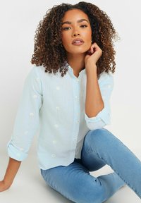 Light blue button-up shirt with small white daisy embroidery, long sleeves rolled up, paired with blue skinny jeans. Smooth texture.
