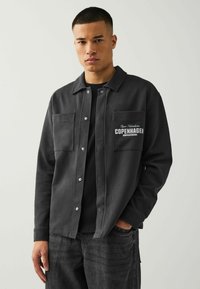 Schwarze Fleece-Hemdjacke mit zwei Vordertaschen, Knopfverschluss und Textdesign. Getragen über einem schwarzen T-Shirt, kombiniert mit dunklen Jeans.