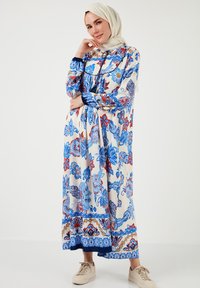 Rochie maxi florală în crem, cu modele albastre și roșii, mâneci lungi și talie strânsă, asortată cu adidași deschiși.