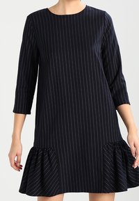 Marineblaues Kleid mit horizontalen weißen Streifen, runder Ausschnitt, Dreiviertelärmel und rüschengesäumtem Saum am unteren Ende.