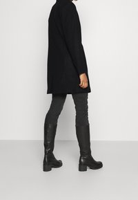 Manteau en laine noire à coupe ajustée, manches longues et fente latérale, associé à un jean skinny gris foncé et des bottes en cuir noires montantes jusqu'au genou.