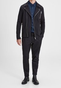 Czarna skórzana kurtka motocyklowa z srebrnymi suwakami, zestawiona z ciemnoniebieską koszulą zapinaną na guziki i czarnymi zwężanymi spodniami. Całość dopełniają czarne botki za kostkę.