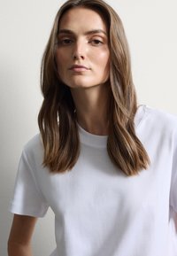 Camiseta blanca de algodón con mangas cortas, cuello redondo, textura suave, llevada por una persona con cabello largo y liso de color castaño frente a un fondo liso.
