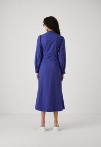 Abito a maniche lunghe, lungo fino al ginocchio, in vivace lino blu con polsini abbottonati e dettagli ricamati sulle maniche, abbinato a scarpe bianche con tacco blocco.