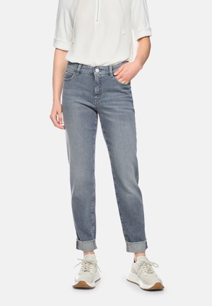 Person trägt hellgraue, umgeschlagene Jeans, weißes Oberteil mit hochgekrempelten Ärmeln und weiße Sneakers, steht mit einer Hand in der Hosentasche.