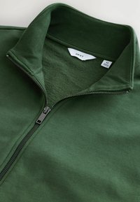 Veste en polaire vert foncé avec un col zippé. Présente une texture douce, une construction à coutures plates et une étiquette avec les informations de taille.