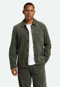 Olivgrüne Cord-Jacke mit durchgehendem Knopfverschluss, schmalem Kragen und einer Brusttasche, kombiniert mit passenden olivfarbenen Hosen und einem weißen Hemd.