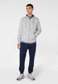 Młody mężczyzna stoi z rękami w kieszeniach, ubrany w szary rozpinany hoodies, niebieską koszulę, granatowe dresy i białe trampki na białym tle.