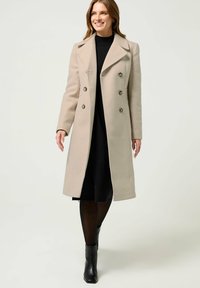 Beige wollen blend jas met een double-breasted ontwerp, grote knopen, een reverskraag en een getailleerde silhouet, gecombineerd met een zwarte jurk en laarzen.