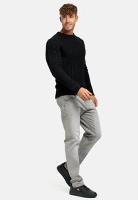 Schwarzer Kabelstrickpullover mit Rundhalsausschnitt, kombiniert mit hellgrauen Slim-Fit-Jeans und schwarzen Sneakers, die ein strukturiertes Muster aufweisen.