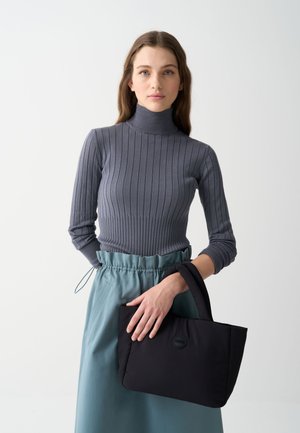 Top gri cu guler înalt ribizat, asortat cu o fustă turcoaz. Modelul ține în mână o geantă de umăr din material textil negru, cu un logo și un design căptușit.