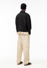 Homme de dos portant une veste noire, un pantalon beige à jambes larges avec poches arrière, et des baskets noires sur un fond blanc.