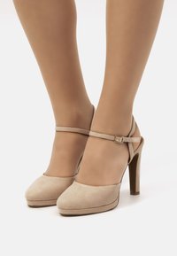 Zapatos de tacón alto de ante beige con plataforma, correa para el tobillo y detalle de hebilla, con una textura suave y un diseño de punta redondeada.