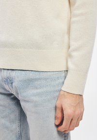 Krēmkrāsas adīts džemperis ar ribotām aprocēm un apakšu, kombinēts ar gaiši ziliem džinsiem. Pie apakšas ir neliels reljefa logotips.