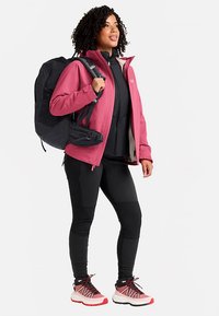Roze softshell jas, zwarte sportlegging en sneakers met roze accenten. Draagt een zwarte rugzak, gestyled voor buitenactiviteiten.