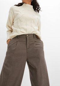 Maglione a collo alto in maglia a treccia crema abbinato a pantaloni larghi in motivo houndstooth marrone, con tasche frontali e chiusura a bottone.