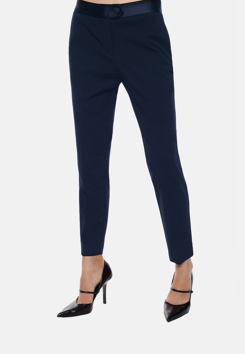 Pantaloni navy con un design aderente, gambe affusolate e dettagli lucidi in vita. Abbinati a décolleté nere a punta e cinturini alla caviglia.