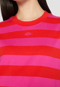 Femme portant un t-shirt rayé horizontal rouge et rose avec un col rond et un petit logo brodé au niveau de la poitrine.