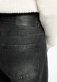 Vaqueros de mezclilla negra con un acabado texturizado, que cuentan con una etiqueta de cuero marcada "EIGHTYFIVE" en la cinturilla y en un bolsillo trasero.