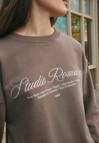 Osoba oblečená v hnědém svetru s nápisem "Studio Romance" a vyšitými názvy měst, včetně New Yorku, Tokia a Londýna, na přední straně.