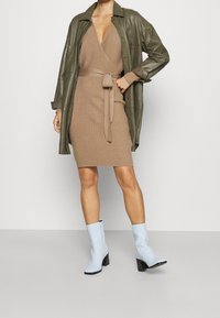 Veste-chemise en cuir kaki portée sur une robe moulante en maille marron avec ceinture nouée, assortie à des bottines bleu clair à talon bloc.