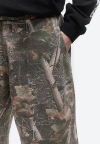 Primo piano di pantaloni con motivo mimetico forestale, che mostrano una mano infilata nella tasca destra e parte di una manica di felpa nera.