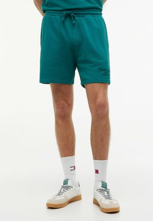 Man draagt teal kleurige shorts met koord, witte sokken met merklogo en witte sneakers met rubberen zolen, staand tegen een witte achtergrond.