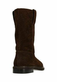 Botte en suède marron jusqu'au genou avec un bout arrondi, texture lisse et semelle plate. Présente des détails de couture le long du dos et des languettes latérales.