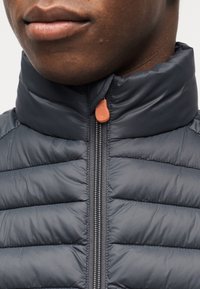 Κοντινό πλάνο ενός ατόμου που φοράει σκούρο γκρι, επενδυμένο μπουφάν με φερμουάρ, ψηλό γιακά και καφέ τράβηγμα φερμουάρ.