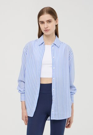 Camicia a righe blu e bianche con bottoni, maniche lunghe, vestibilità rilassata e tessuto morbido, indossata sopra un crop top bianco e leggings blu scuro.