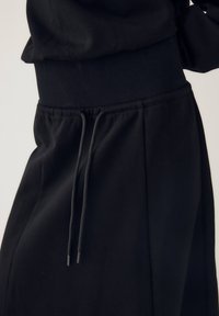 Pantalon de survêtement noir taille haute avec une ceinture élastique et des cordons noirs, porté avec un haut noir assorti.