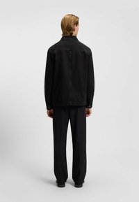 Zwarte denimjas met een puntkraag, met knoopsluiting bij de mouwen en een relaxte pasvorm, gecombineerd met zwarte broek, gezien van achteren.