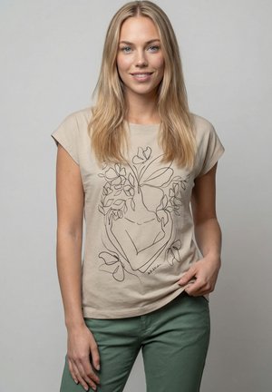 Junge Frau mit langen blonden Haaren, die ein beiges T-Shirt trägt, auf dem ein schwarzes Linienkunst-Design einer gesichtslosen Figur mit Blättern und Blumen abgebildet ist.