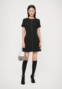 Černé šaty s krátkými rukávy a přímým střihem, zdobené kovovými cvočky podél středu. Kombinovány s podkolenkami po kolena a malou texturovanou kabelkou.