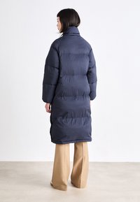 Jūras zila pūkainā jaka ar augstu apkakli un sānu rāvējslēdzējiem, ar quilted dizainu un garām piedurknēm, valkāta virs beigtu platām biksēm.