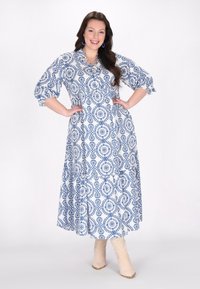 Rochie maxi albă cu model circular albastru, mâneci umflate, guler și închidere cu nasturi. Asortată cu cizme cu gleznă de un bej deschis.