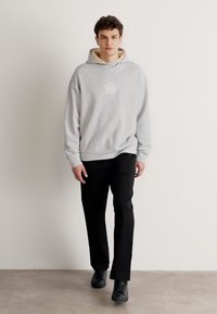 Šedá mikina s kapucí a předním logem, doplněná černými kalhotami a černými botami, na modelu na jednobarevném pozadí.