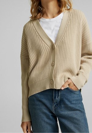 Beige gebreide cardigan met een V-hals, geribbelde textuur en drie knopen. Draag het over een wit T-shirt, gecombineerd met blauwe jeans.