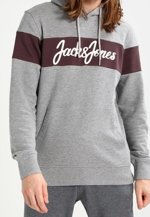 Osoba ubrana w szarą bluzę z kapturem z bordowym paskiem na piersi i białym logo "Jack & Jones", w połączeniu z szarymi spodniami.