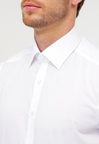 Hvit skjorte med klassisk krage, laget av glatt stoff, med en frontlomme med knapper og en teksturert overflate.