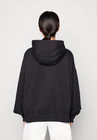 Svart oversized hoodie i bomullsmix, med en stor huva, sänkta axlar och ribbad nederkant. Bakifrån, slät textur.