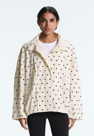 Nainen pukeutune oversize-koon kermaan mustilla pilkuilla pilkulliseen takkiin ja mustiin housuihin, seisoo yksinkertaisen valkoisen taustan edessä.