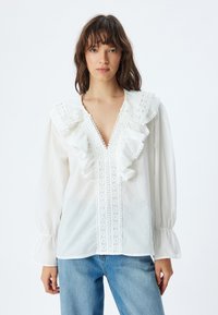 Witte blouse met ruffles, kantafwerking aan de voorkant en ballonsleeves. Stof lijkt lichtgewicht en textuur te hebben.