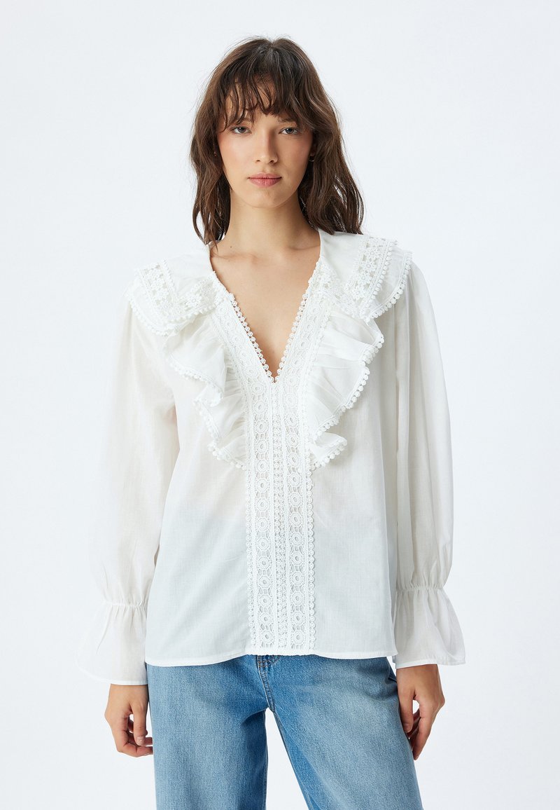Witte blouse met ruffles, kantafwerking aan de voorkant en ballonsleeves. Stof lijkt lichtgewicht en textuur te hebben.