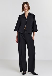 Donna in piedi con pantaloni neri a gamba larga con fenditure anteriori, camicia abbottonata larga abbinata e tacchi a punta neri su uno sfondo uniforme.